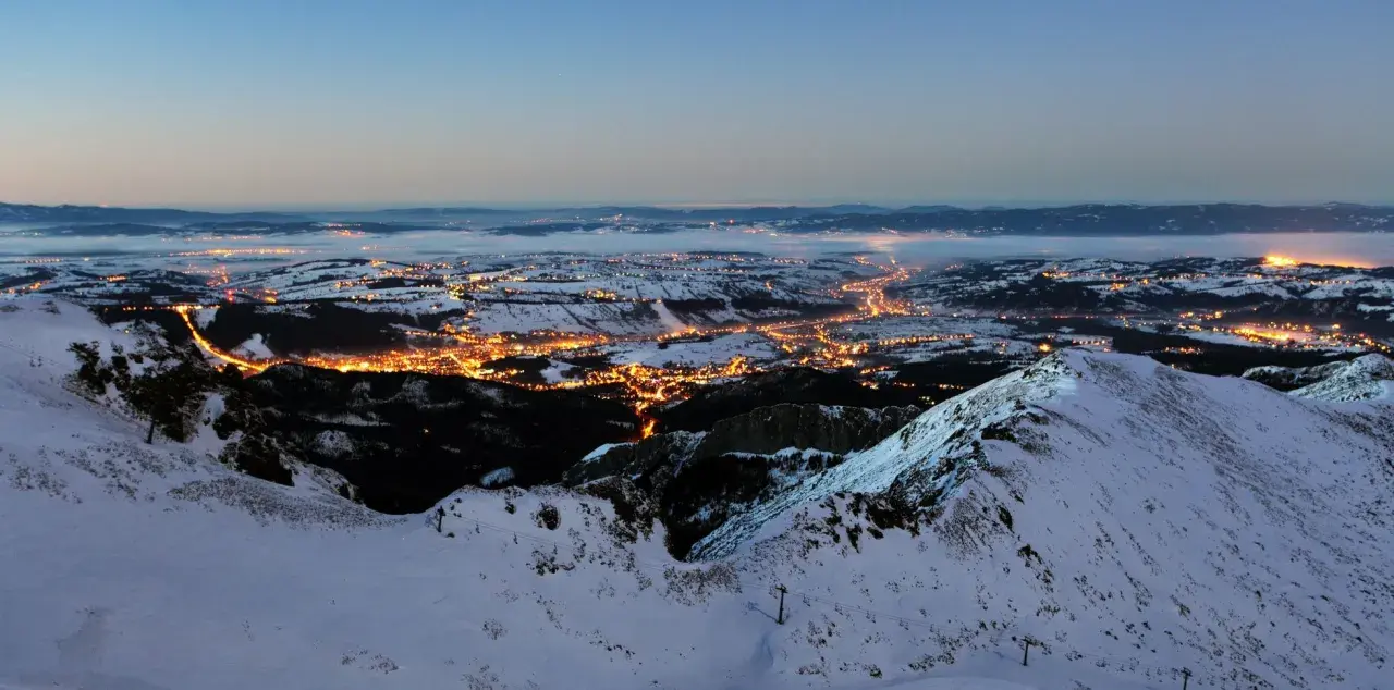 Widok z góry na oświetlone miasto w dolinie, otoczone ośnieżonymi górami. Tak wygląda nocą miejsce, gdzie leży Zakopane.