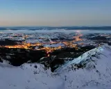 Widok z góry na oświetlone miasto w dolinie, otoczone ośnieżonymi górami. Tak wygląda nocą miejsce, gdzie leży Zakopane.