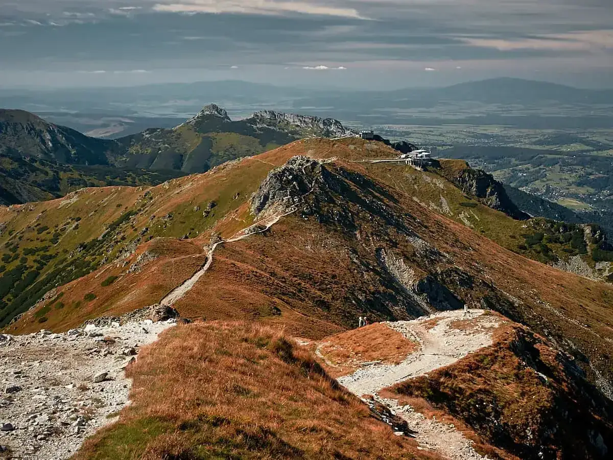 Szlak na Giewont, świnica poziom trudności. Kręta ścieżka wśród traw i skał, w oddali widać budynki na szczycie.